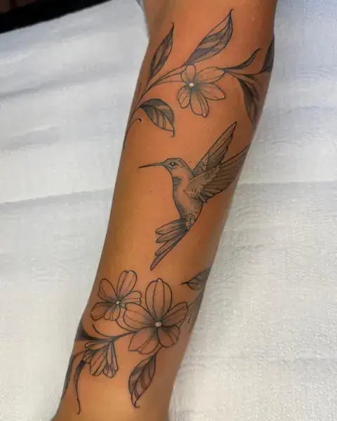 Tatuagem fine line de beija-flor com flores no antebraço